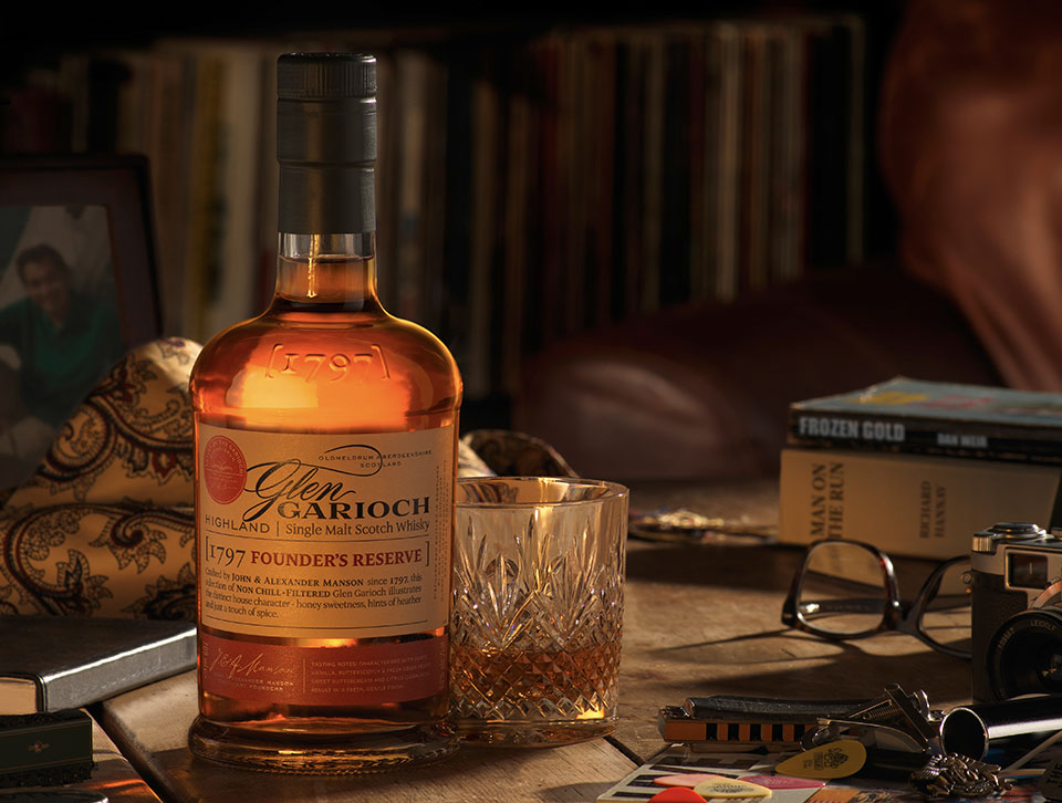 A bottle and glass of neat Glen Garioch Whiskey on a table with glasses, books and keys