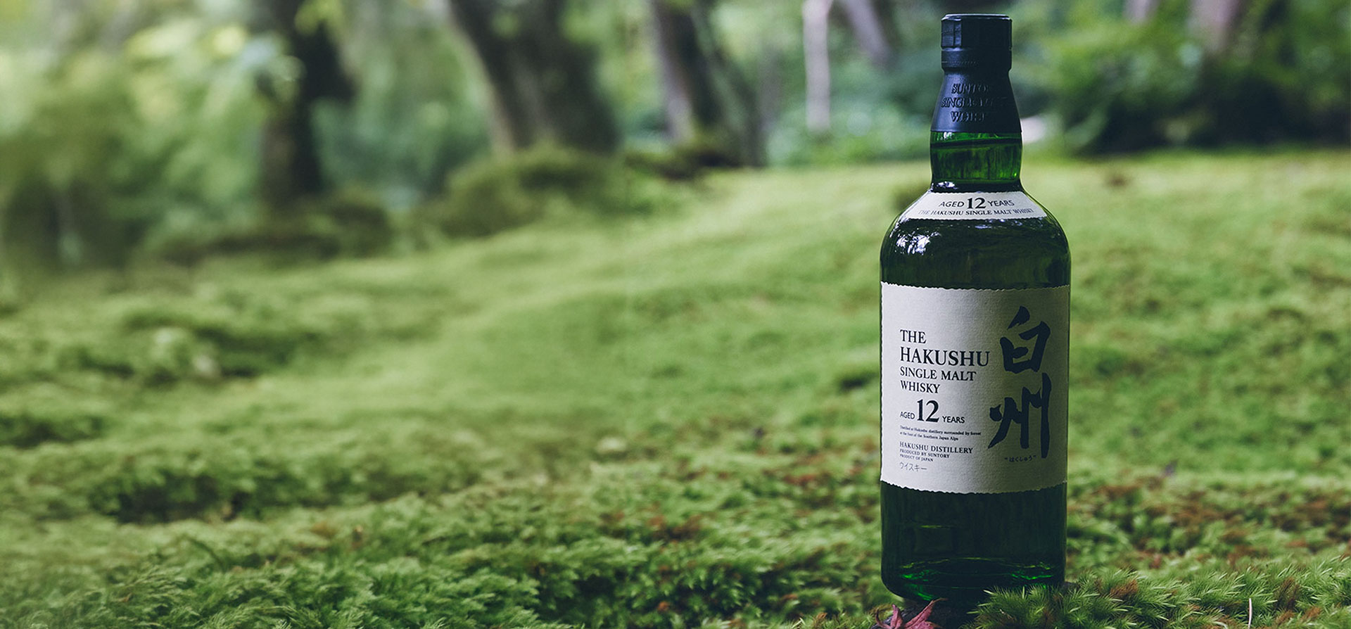 A bottle of Hakushu whisky placed on grass in an outdoor forest setting.