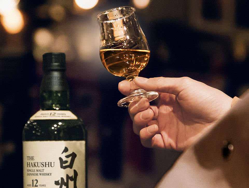 A hand holding a neat glass of Hakushu whisky next to its bottle