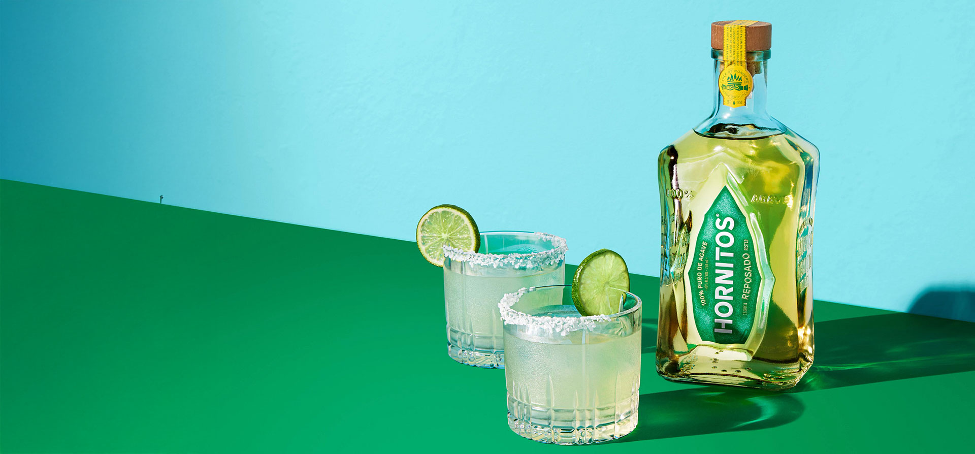 A bottle of Hornitos and 2 glasses of cocktails on a light blue and green duo tone background