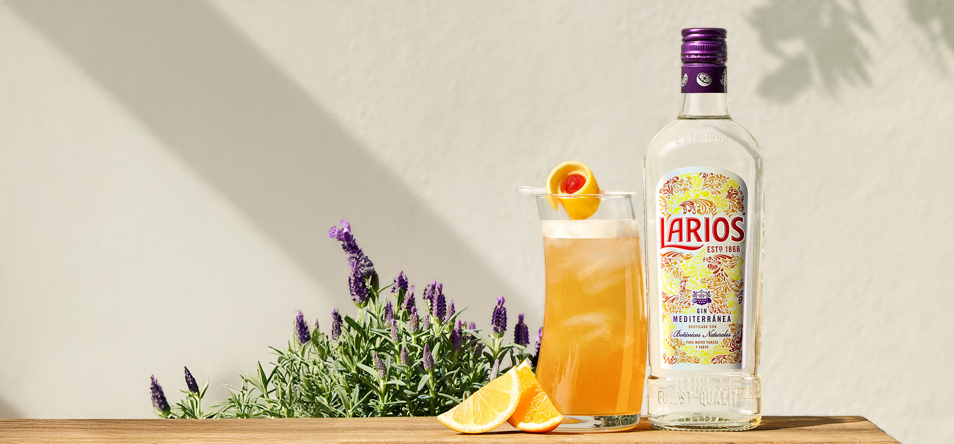 A bottle and glass of Larios against a white wall and lavender flowers