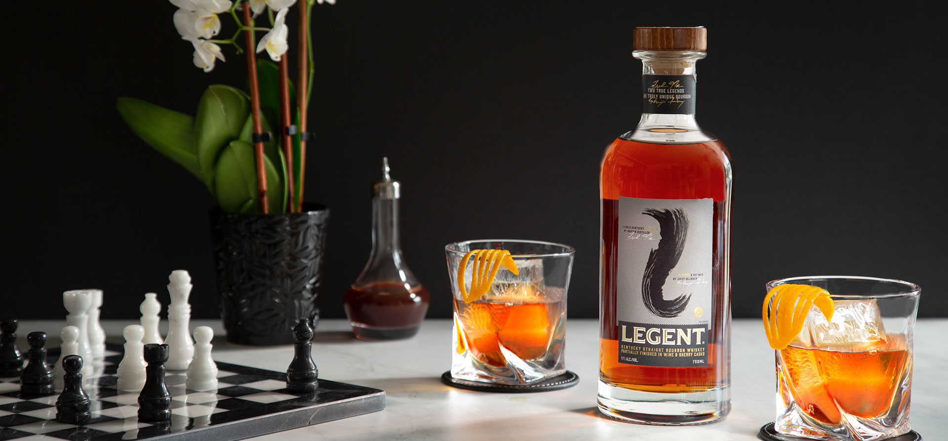 A bottle of Legent whiskey and glasses on a table set with a chessboard.