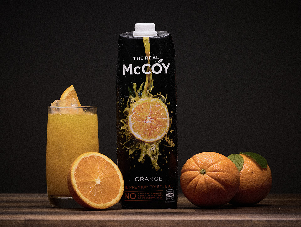 The Real McCoy carton, glass of juice and whole oranges on a wooden table