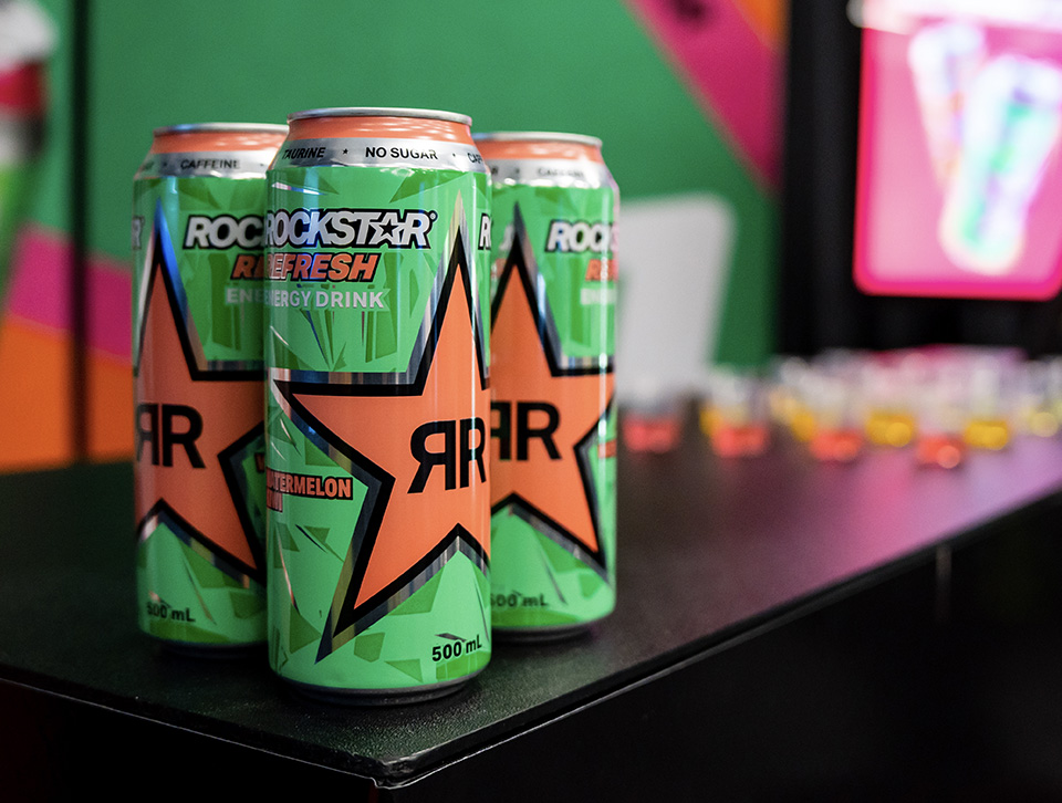 Three cans of green Rockstar with cups of drinks in the background