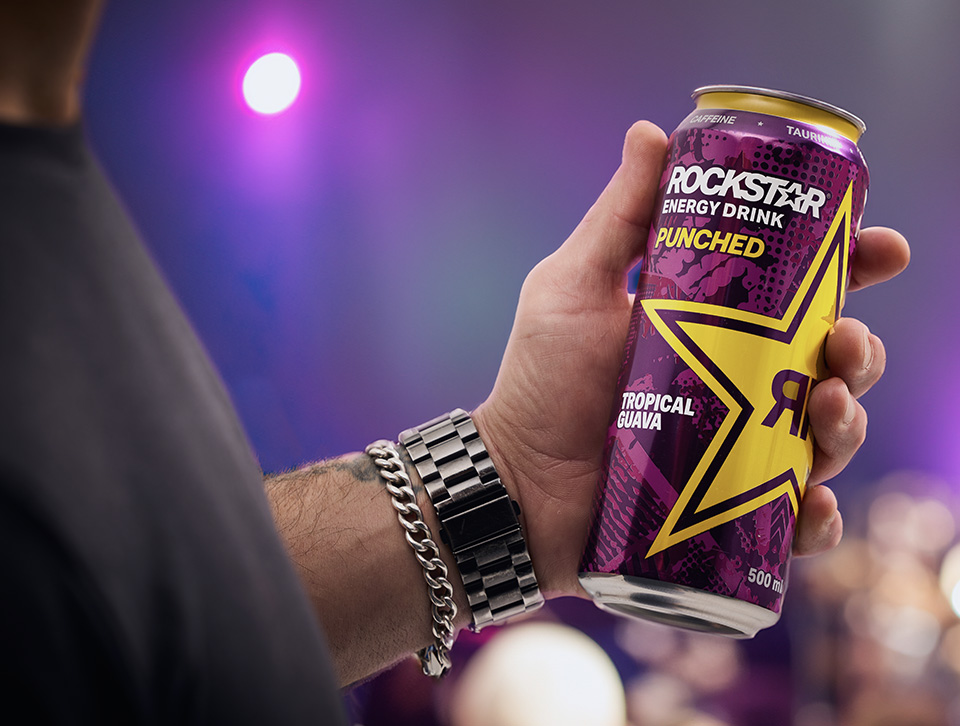 A hand holding a can of Rockstar Tropical Guava against a purple background