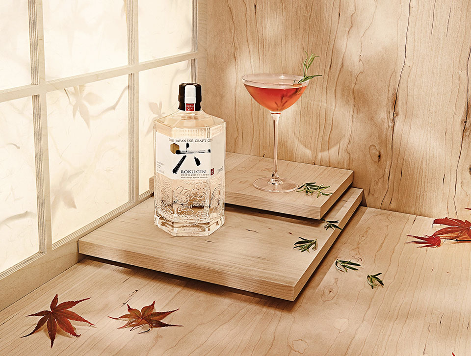 A bottle of Roku and a cocktail on a wooden platform with rosemary and maple leaves