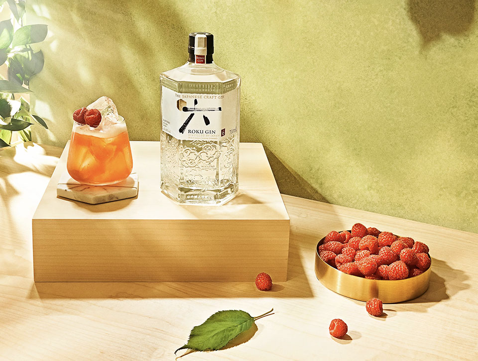 A bottle of Roku and a cocktail on a wooden platform with raspberries 