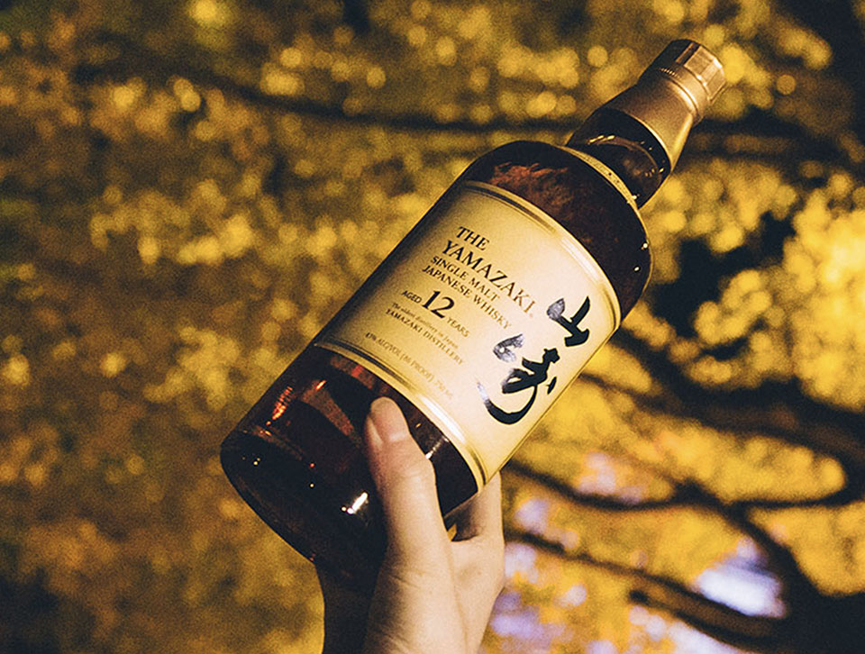 A hand holding a bottle of Yamazaki whisky with a tree in the background.