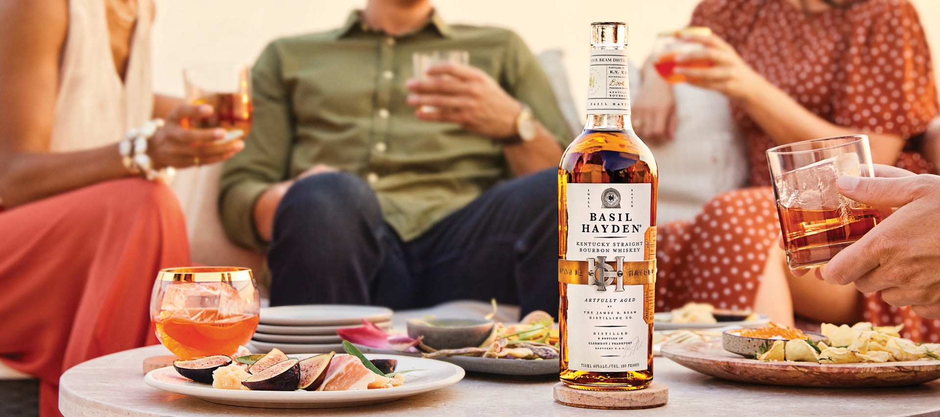 People sitting around a table of party food and a bottle of Basil Hayden