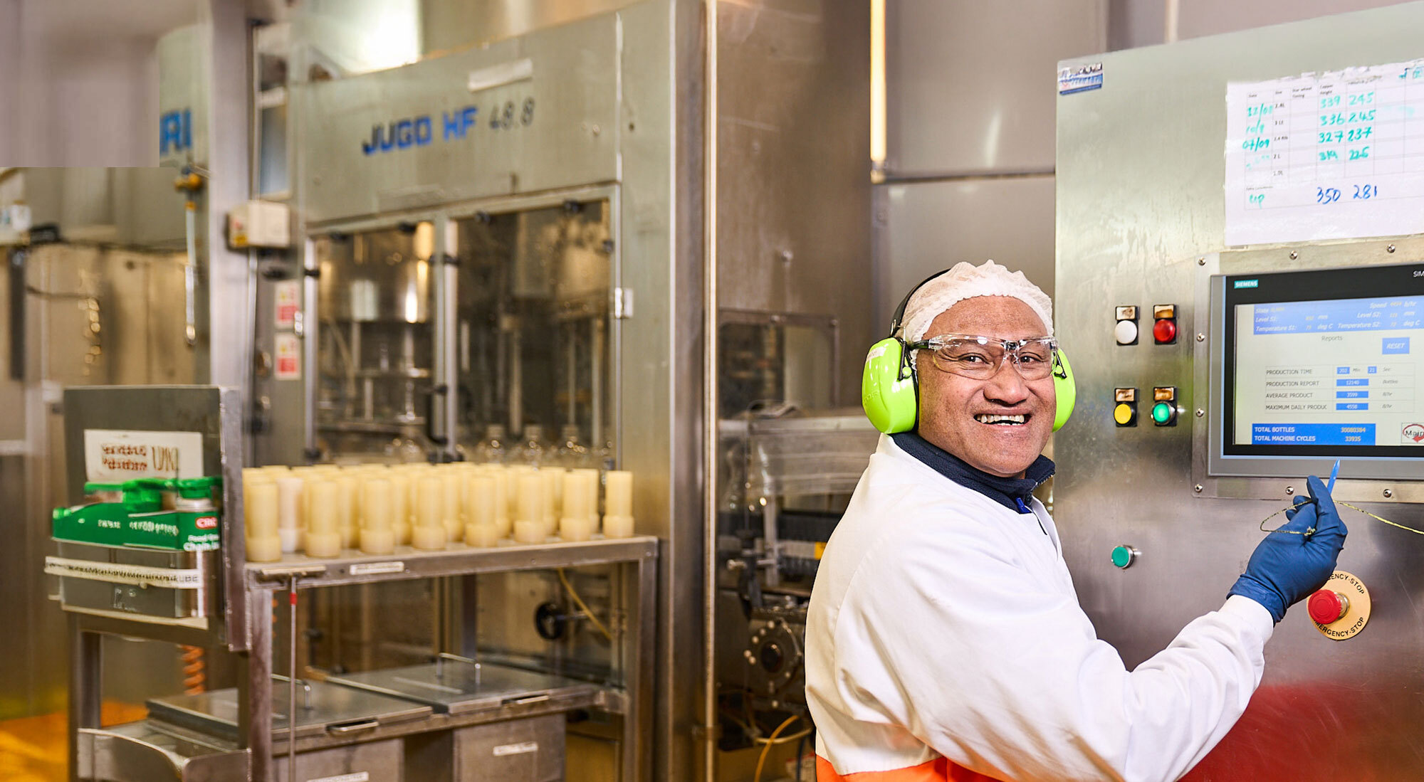 A factory worker in earmuffs and hairnet using a machine with monitor.