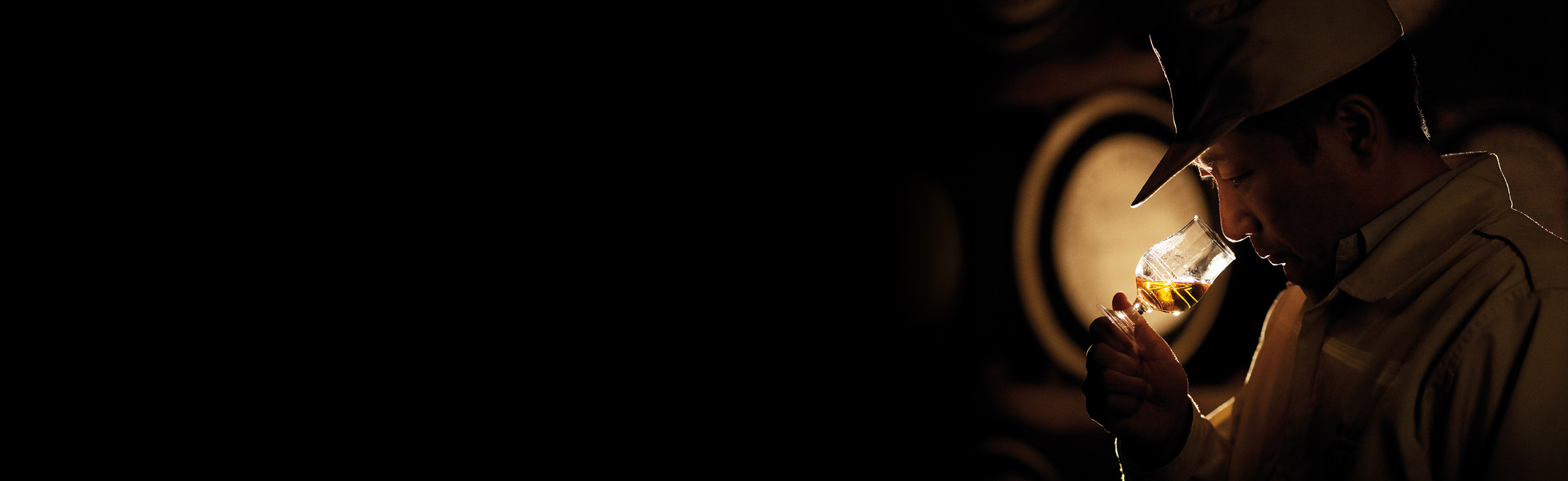 A profile shot of a man standing in front of alcohol barrels, nosing a glass of whisky.