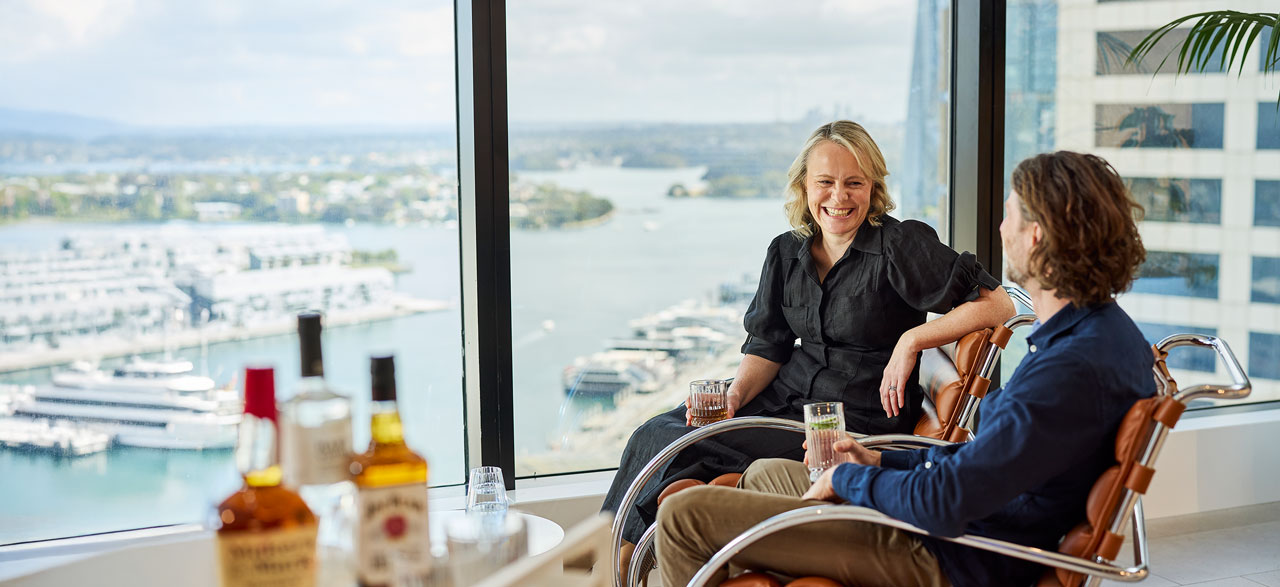 Smiling woman and man sitting in casual leather armchairs holding drinks next to a table with three Suntory Alcohol alcohol spirits. They sit beside a large window that overlooks Sydney Harbour.