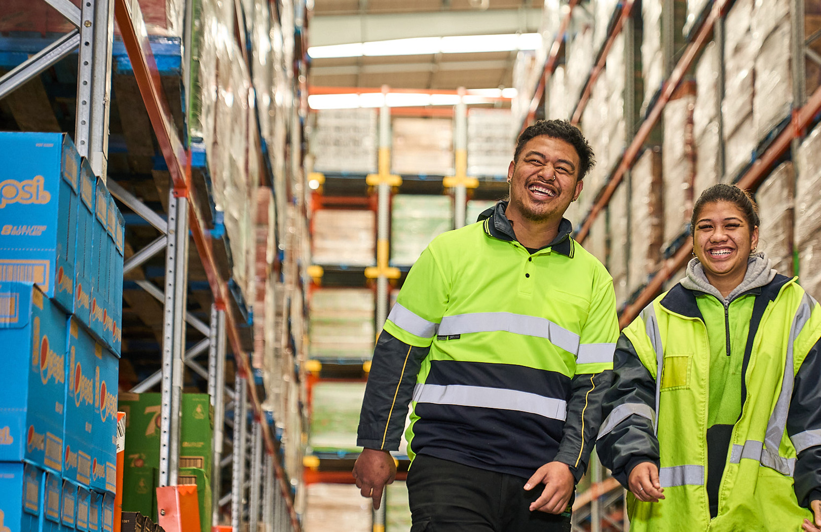 Two workers wearing high-vis vests walking through a warehouse with big smiles