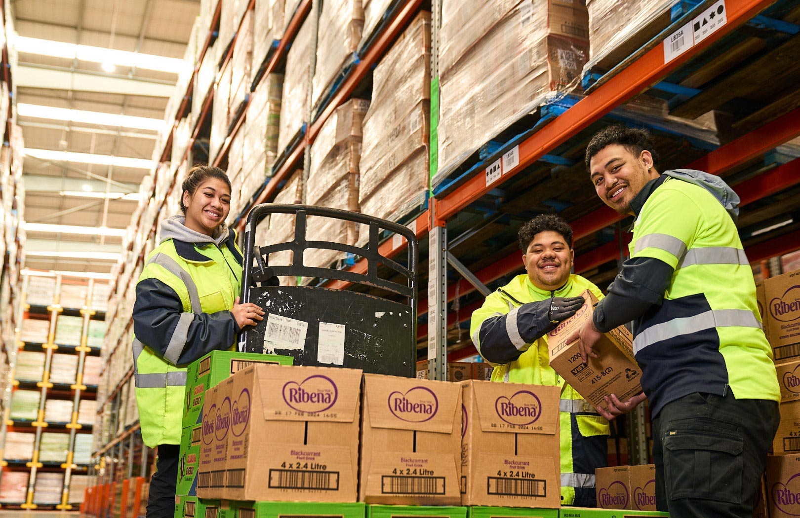 Two workers wearing high-vis vests holding boxes in a warehouse and posing for camera