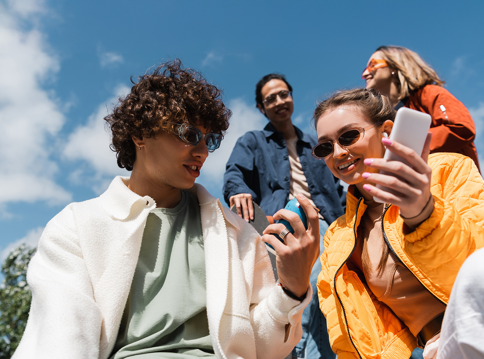 A group of young people outdoors, holding their phone and a can of drink