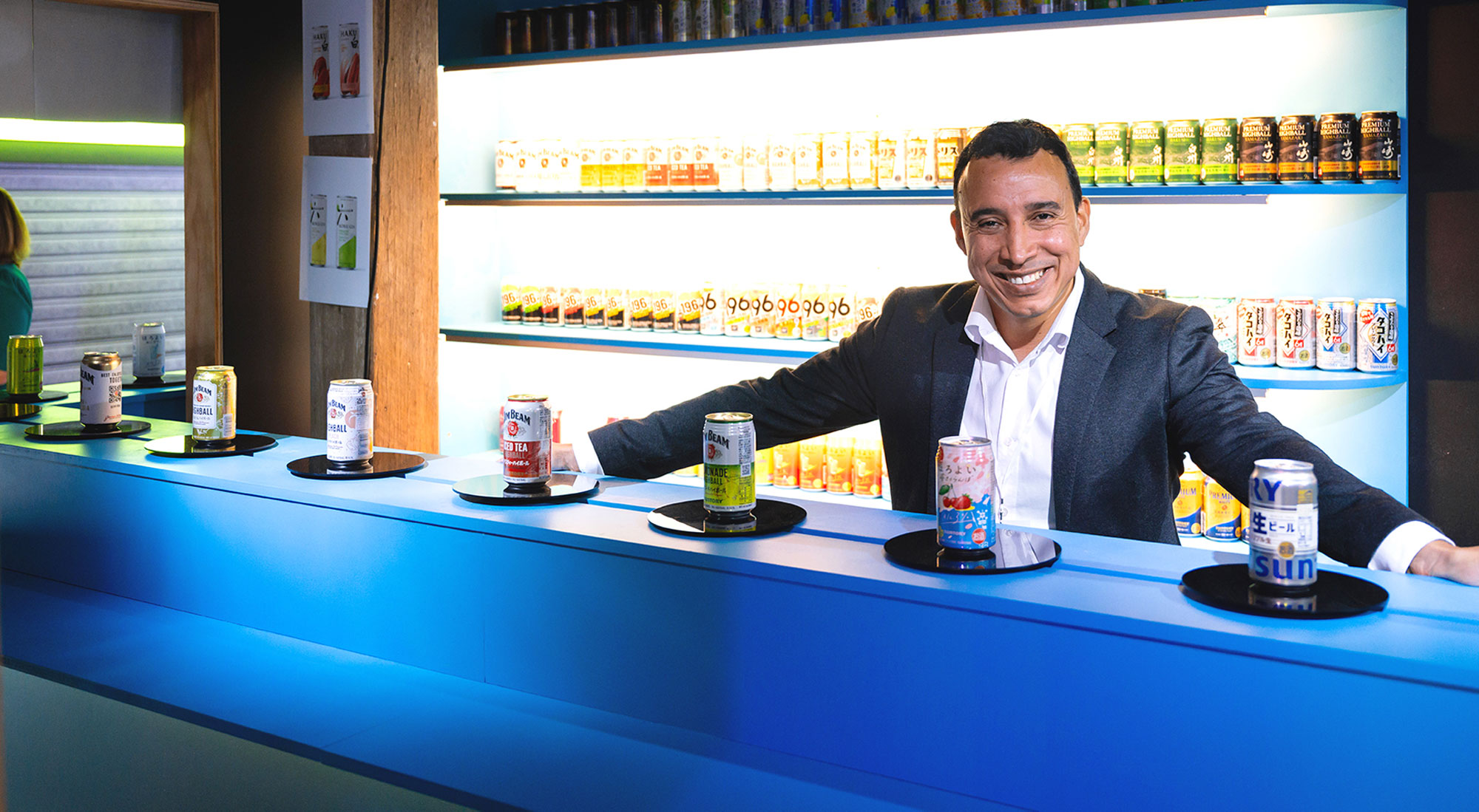 Man in suit standing at the dark blue bar counter with a line up of various Suntory alcoholic brand cans