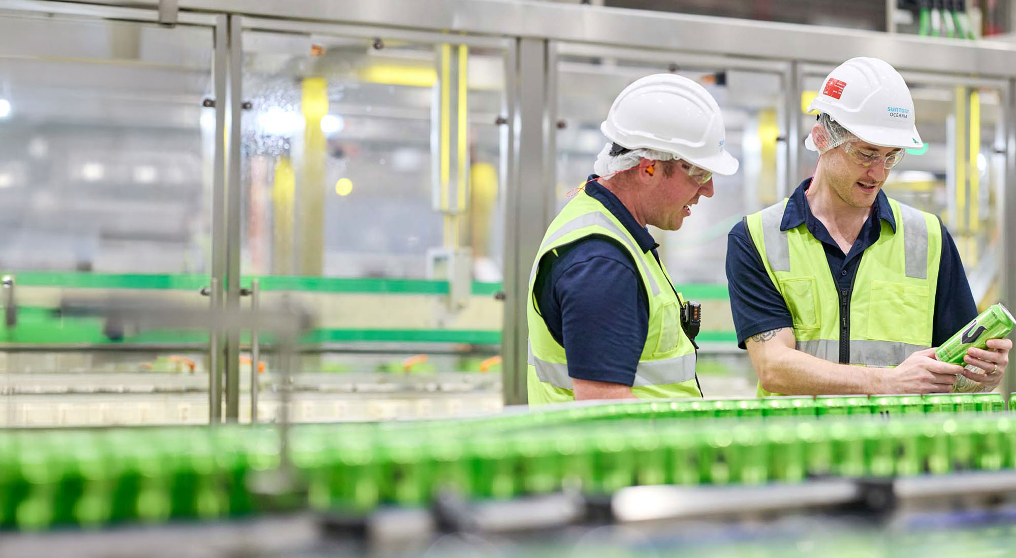 Two men in high-vis vests in a factory with V Energy cans across the production line