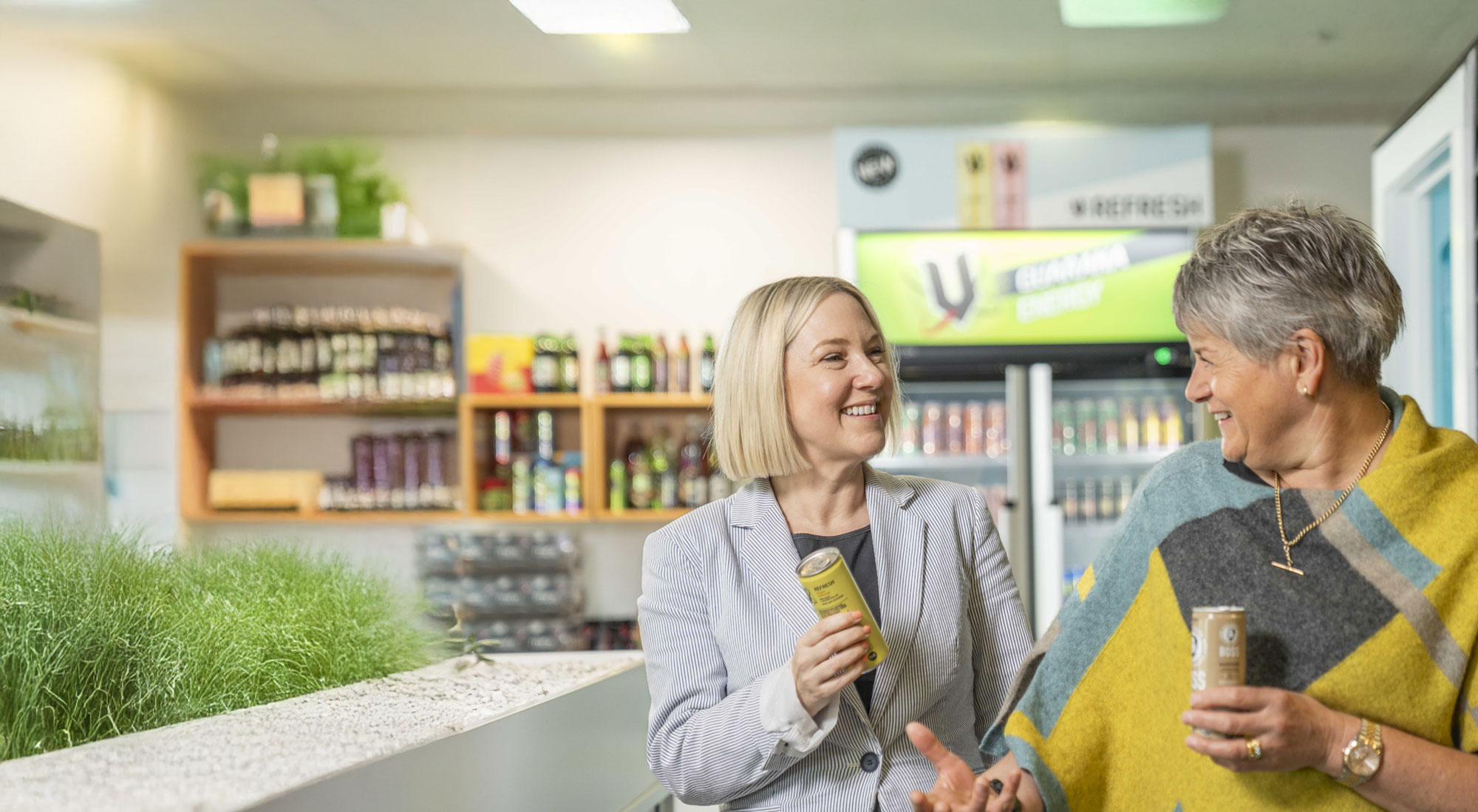 Two women in a cafeteria smiling with a can of V Energy and Boss Coffee in their hands.